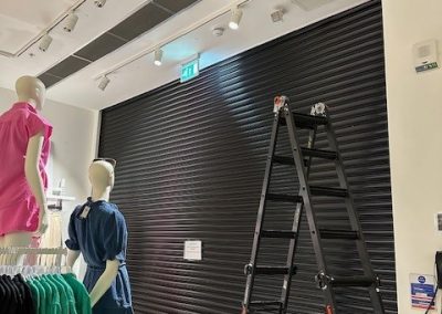 Ladder set up beside black roller shutter door under repair inside NEXT store
